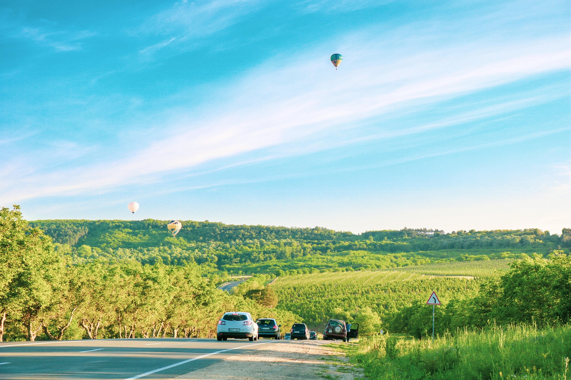 Autos fahren eine Landstraße entlang, umgeben von Natur und Weinbergen. Über der Szenerie steigen mehrere bemannte Heißluftballons in den Himmel. Moldau bietet vor allem Wein und Natur.