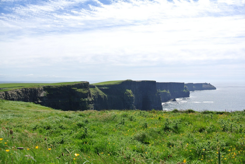 Steile Küstenklippen in Nordirland mit grünen Wiesen, schroffen Felsformationen und dem Meer, das unterhalb gegen die Felsen brandet.
