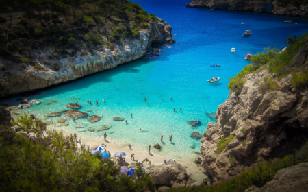 Heller Sandstrand auf Mallorca mit türkisblauem, klarem Meer und sanftem Wellengang, eingerahmt von Felsen und Pinien unter blauem Himmel.