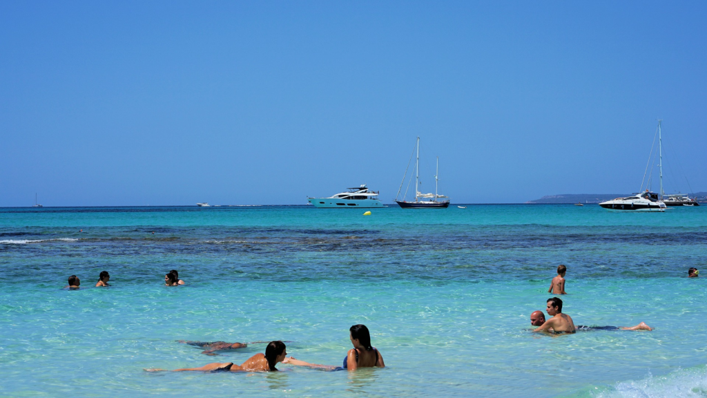 Mallorca‑Strand mit Badegästen, kristallklarem türkisblauem Meer und Yachten in der Bucht.