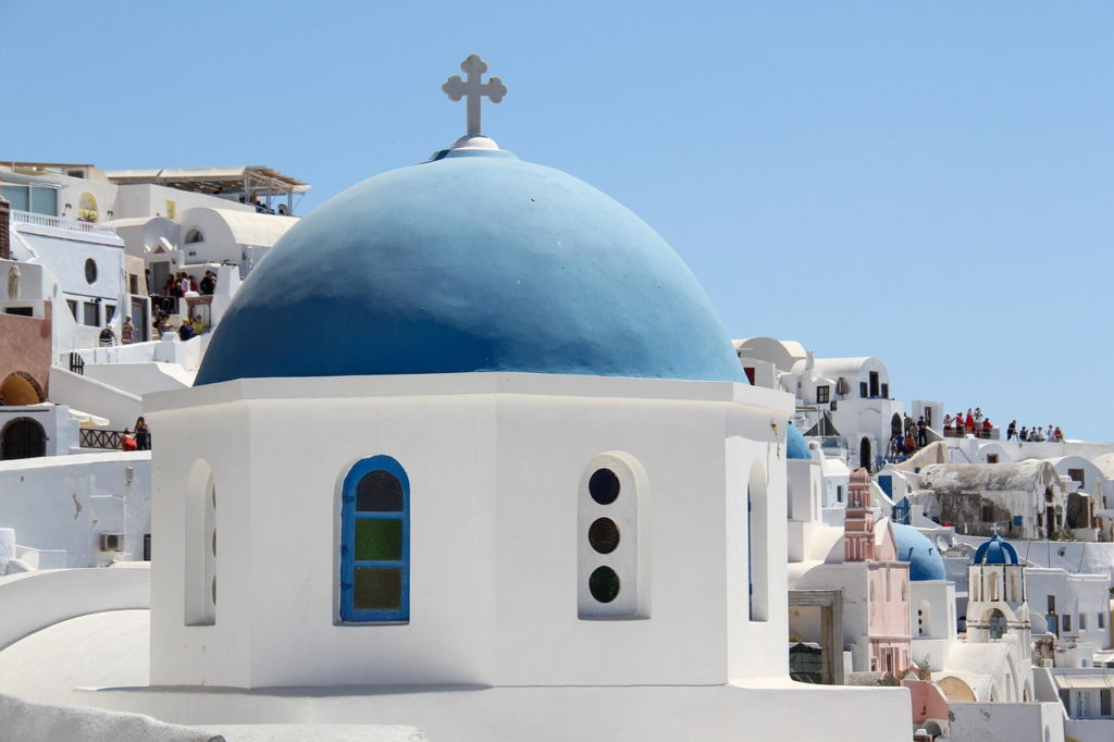 Weiße Kirche mit blauer Kuppel auf einer Kykladen‑Insel, umgeben von traditionellen Häusern und Blick auf das Meer.